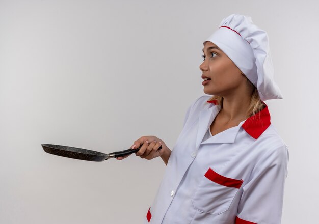 Standing in profile view surprised young cook female wearing chef uniform holding out frying pan to side with copy space