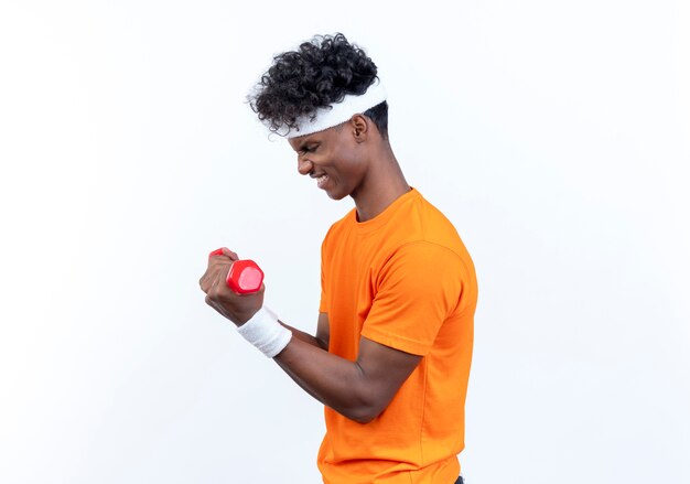 Standing in profile view tense young afro-american sporty man wearing headband and wristband exercising with dumbbell