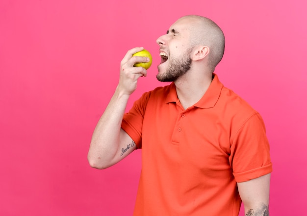 Free photo standing in profile view with closed eyes young sporty man trying bites of apple isolated on pink wall