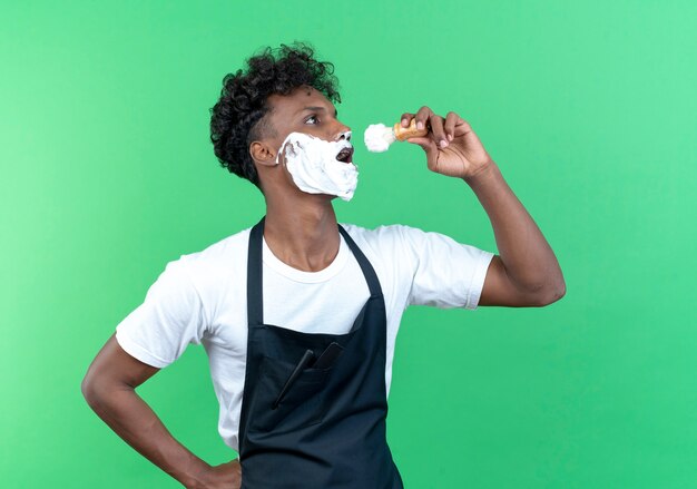 Standing in profile view young afro-american male barber wearing uniform with shaving cream applied on his face holding shaving brush and singing