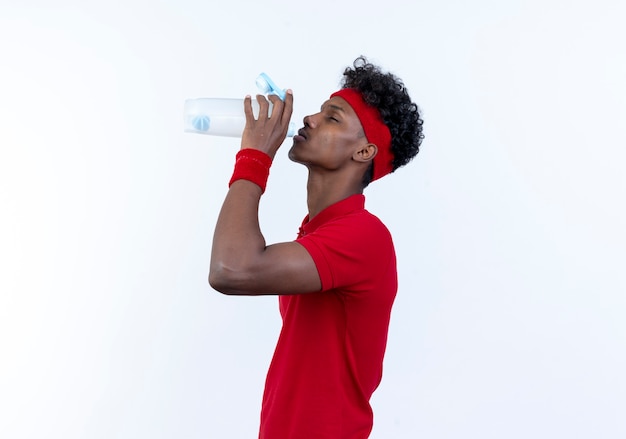 Standing in profile view young afro-american sporty man wearing headband and wristband drinking water from bottle isolated on white