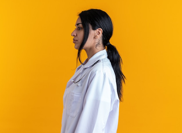 Standing in profile view young female doctor wearing medical robe with stethoscope isolated on yellow background