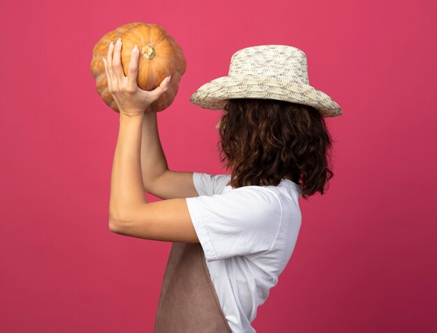 Standing in profile view young female gardener in uniform wearing gardening hat raising pumpkin