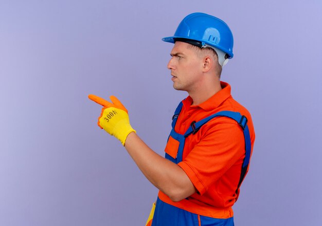 Standing in profile view young male builder wearing uniform and safety helmet in gloves point at side  on purple