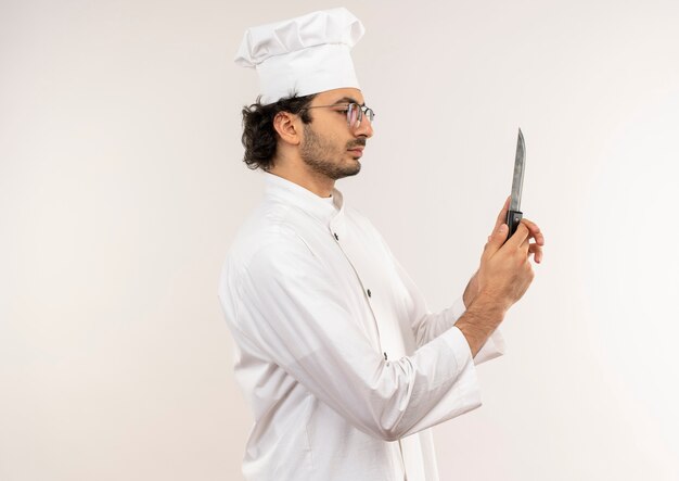 Standing in profile view young male cook wearing chef uniform and glasses holding and looking at knife isolated on white wall