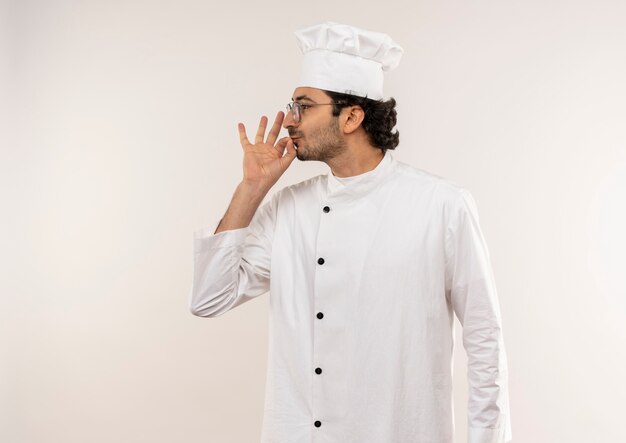 프로필보기 젊은 남성 요리사 입고 요리사 유니폼과 흰 벽에 고립 된 맛있는 제스처를 보여주는 안경에 서