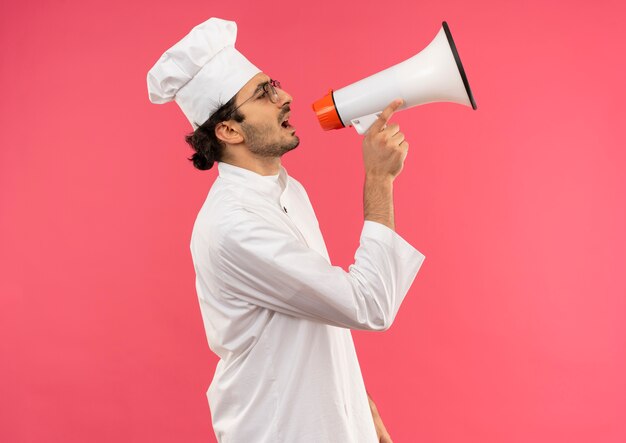 Стоя в профиль, молодой мужчина-повар в униформе шеф-повара и в очках говорит по громкоговорителю, изолированному на розовой стене