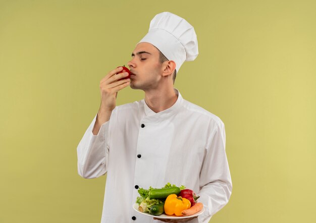 Стоя в профиль, молодой мужчина-повар в униформе шеф-повара держит овощи на тарелке и нюхает помидоры