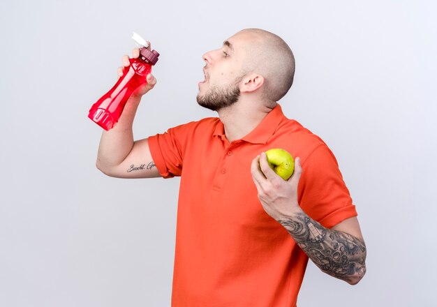 縦断ビューに立って白い壁に分離された水のボトルとリンゴを保持している若いスポーティな男