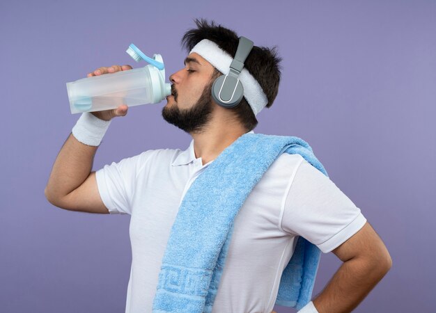 Standing in profile view young sporty man wearing headband and wristband with headphones drinks water with towel on shoulder putting hand on hip isolated on green