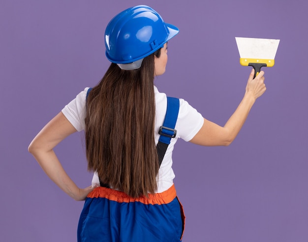 Стоя позади взгляда молодой женщины-строителя в униформе, держащей шпатель, изолированной на фиолетовой стене