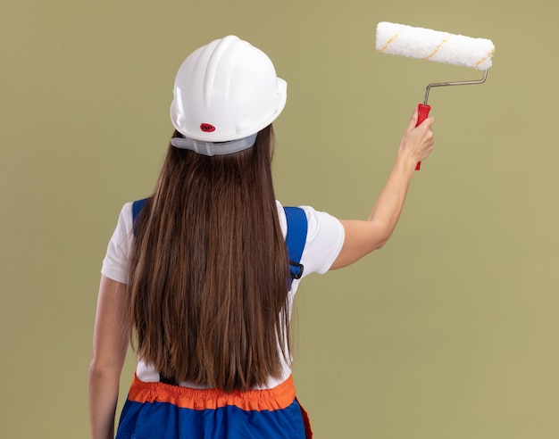 Levandosi in piedi dietro la donna giovane costruttore di vista in uniforme che tiene la spazzola del rullo isolata sulla parete verde oliva