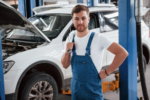 Standing with wrench in hand. Employee in the blue colored uniform works in the automobile salon