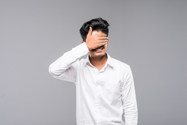 Standing young indian man covering his eyes with his hands, isolated on a white wall.