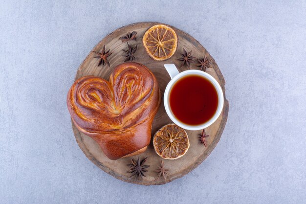 Звездчатый анис, сушеные дольки лимона, чашка чая и сладкая булочка на деревянной доске на мраморной поверхности