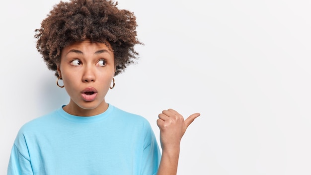 Free photo startled amazed young woman with dark curly hair points thumb away gasps from wonder shows something breathtalking dressed in casual blue t shirt demonstrates amazing offer or advertisement