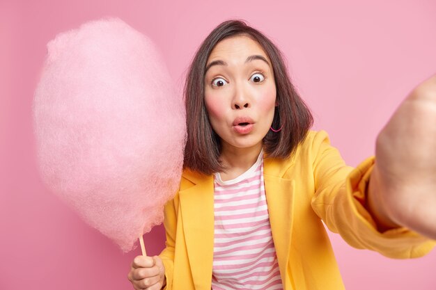  startled Asian woman stares bugged eyes  makes photo of herslef holds tasty cotton candy likes eating something sweet poses against pink wall in stylish clothes