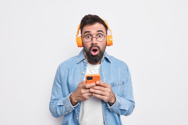 Startled bearded man stares impressed, uses mobile phone and stereo headphones for listening music in playlist wears denim shirt 