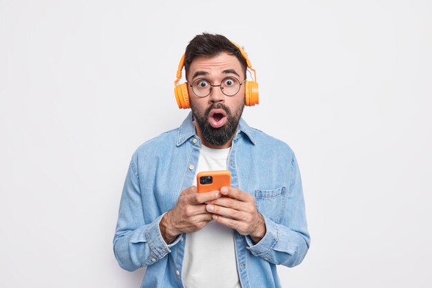 Startled bearded man stares impressed, uses mobile phone and stereo headphones for listening music in playlist wears denim shirt 