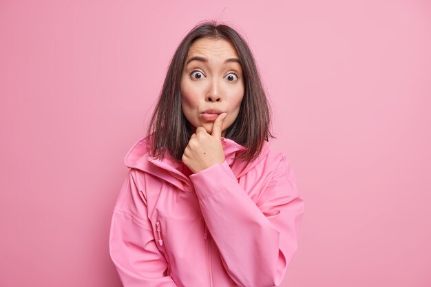 Startled brunette young Asian woman stares surprised  has shocked puzzled expression wears jacket with hood keeps eyes wide opened isolated over pink  wall. Omg concept.