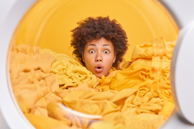 Startled curly haired busy young woman surrounded by yellow laundry loads dirty linen does home chores poses through washing maching door