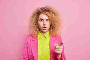 Foto gratuita la donna dai capelli ricci emotiva spaventata si trova senza parole davanti a un'offerta incredibile vestita con una camicia e una giacca luminose isolate sul muro rosa ansimando dalla meraviglia. basta guardare lì