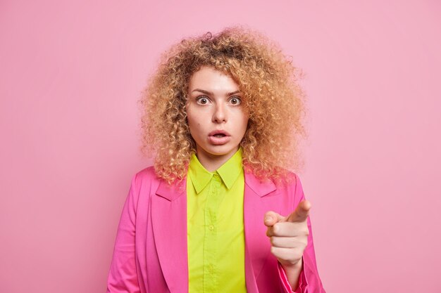 Startled emotional curly haired woman stands speechless points in front sees amazing offer dressed in bright shirt and jacket isolated over pink wall gasps from wonder. Just look there