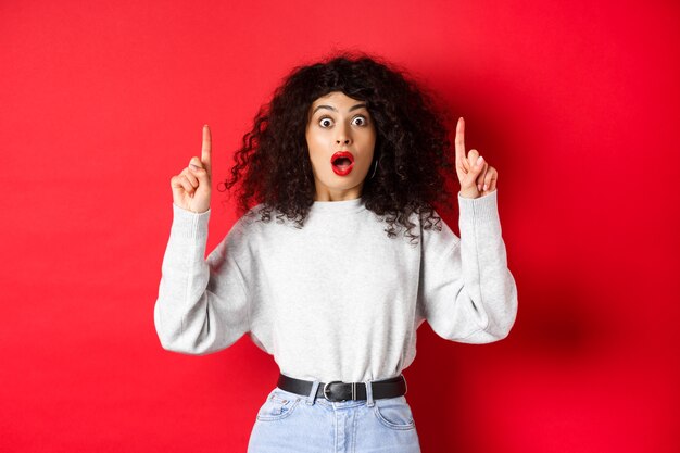 Startled girl showing amazing news drop jaw and gasping surprised as pointing fingers up at empty sp...