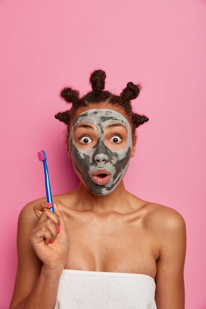 Startled scared woman stares with bugged eyes , holds tooth brush, going to clean teeth after removing beauty mask from face, cares about beauty and hygiene, wears bath towel over naked body