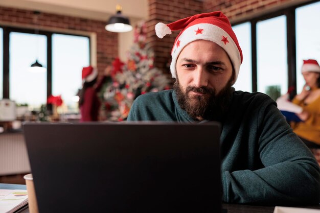冬のシーズン中に会社のプロジェクトに取り組むために、営業所でラップトップを使用するスタートアップの従業員。クリスマス ツリーと休日の装飾と職場でレポートに取り組んでいるサンタ帽子をかぶった男。