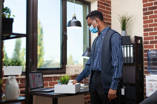 Free photo startup employee wearing covid mask putting personal belongings in tray giving a last look at his desk with laptop. laid off office worker looking with regret at portable computer.