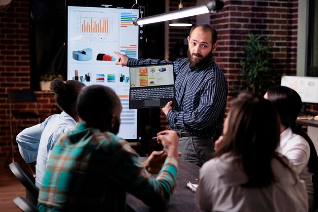 Startup entrepreneur holding laptop with sales presentation charts pointing at wall screen tv in late night meeting. Caucasian man presenting marketing strategy to coworkers working overtime.