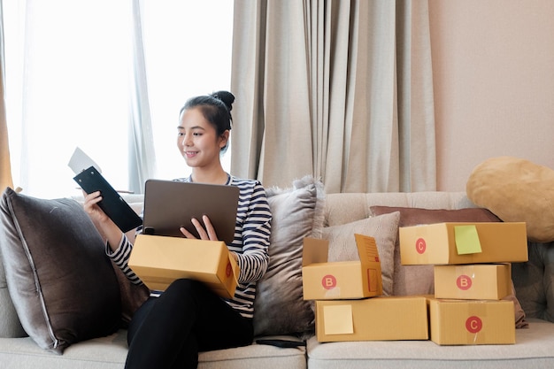 Запуск малого бизнеса, молодая азиатская женщина проверяет онлайн-заказ на цифровом планшете и упаковывает коробки для продуктов для отправки клиентам. работа в домашнем офисе.