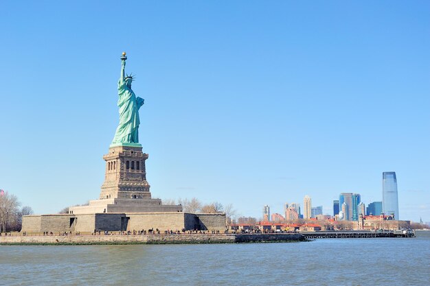 자유의 여신상은 맑고 푸른 하늘이 있는 허드슨 강 위로 고층 빌딩이 있는 뉴욕 맨해튼 시내 스카이라인을 마주하고 있습니다.