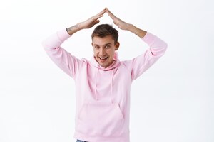 Stay home and be safe portrait of handsome blond guy in pink hoodie showing roof sign hold hands above head encourage people selfdistansing during covid19 pandemic white background