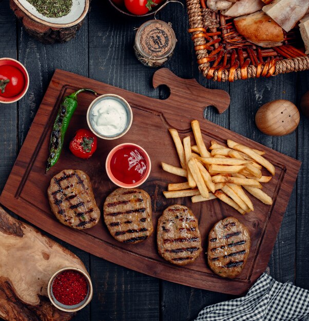 Steak pieces with french fries, grilled pepper and tomato, and sauces on a wooden board.