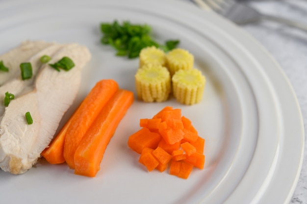 Паровая куриная грудка на белой тарелке с зеленым луком, кукурузой и нарезанной морковью.