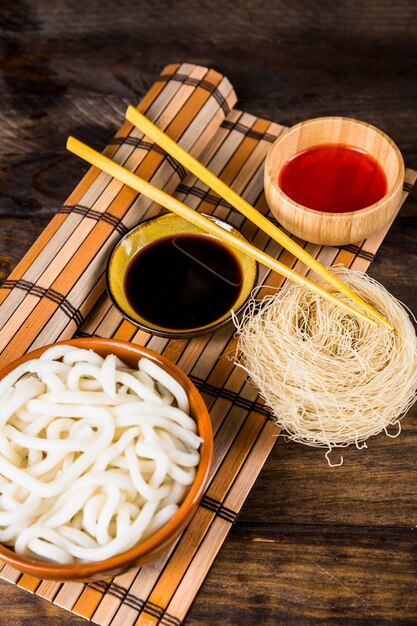 Steams udon noodles; rice vermicelli and sauces with wooden chopsticks over the place mat against wooden table