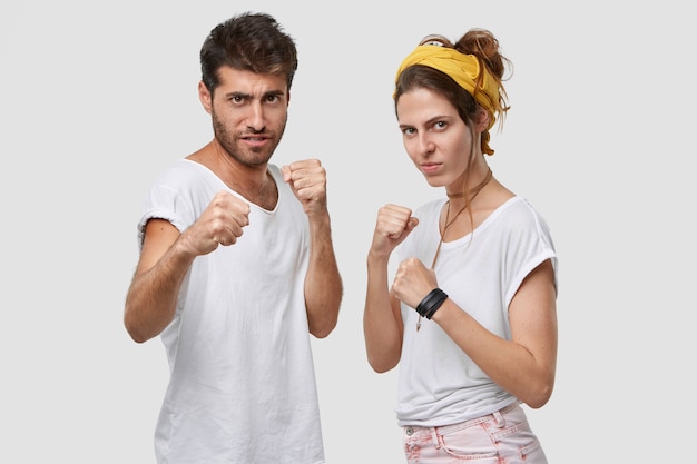 Free photo stern beautiful lady and her unshaven boyfriend stand together, keep hands in defensive gesture, looks seriously, ready to fight and protect themselves, pose against white wall
