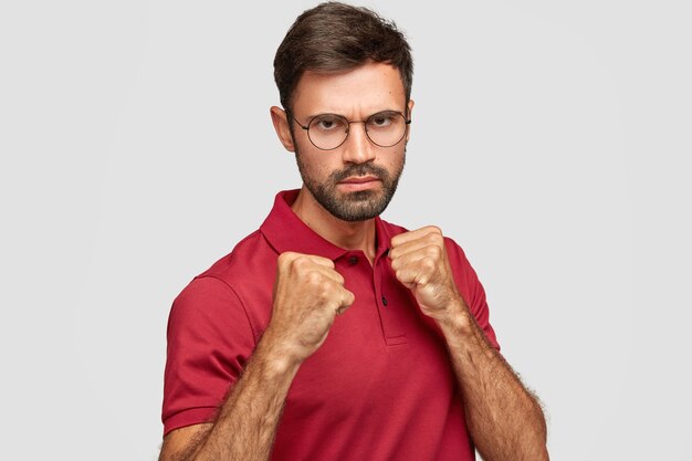 스턴 심각한 형태가 이루어지지 않은 남성은 주먹을 쥐고 경쟁자와 싸울 준비를하고 눈썹 아래를 보며 불쾌한 표정을 지으며 캐주얼 한 빨간 티셔츠를 입고 실내 흰색 벽에 서 있습니다.