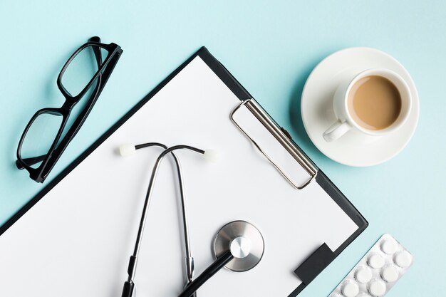 Stethoscope on clipboard with blister pack; spectacles and cup of coffee over desk