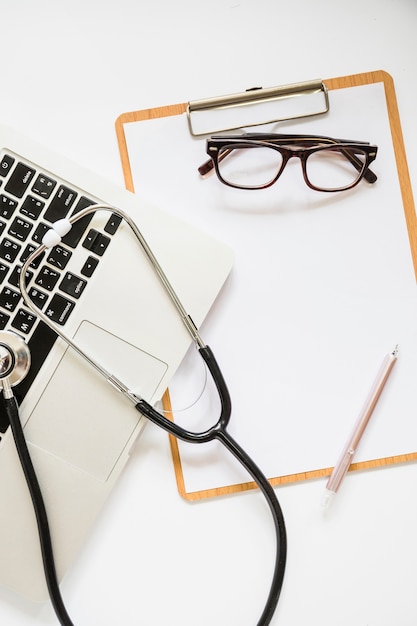 Stethoscope over the laptop and clipboard with eyeglasses and pen on white background