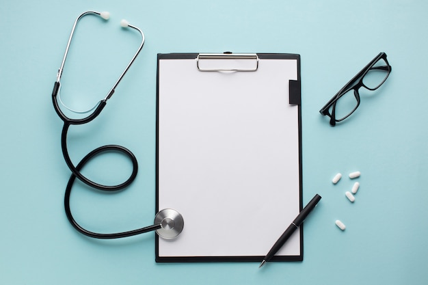 Stethoscope and pen on clipboard near pills with spectacles over blue surface