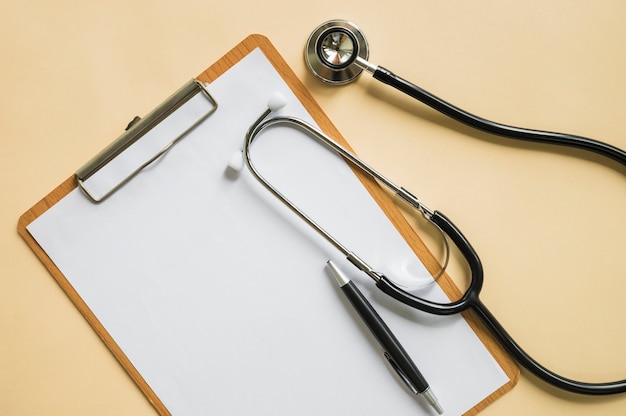 Stethoscope and pen over the clipboard with blank white paper on beige background