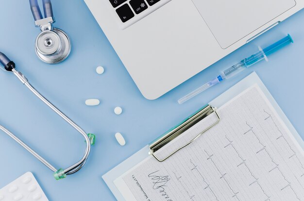 Stethoscope; pills; syringe; laptop and ecg medical report on clipboard over blue backdrop