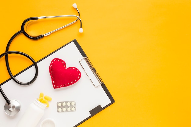 Stethoscope; stitched heart; medicine falling from bottles; blister packed medicine with clipboard over yellow table