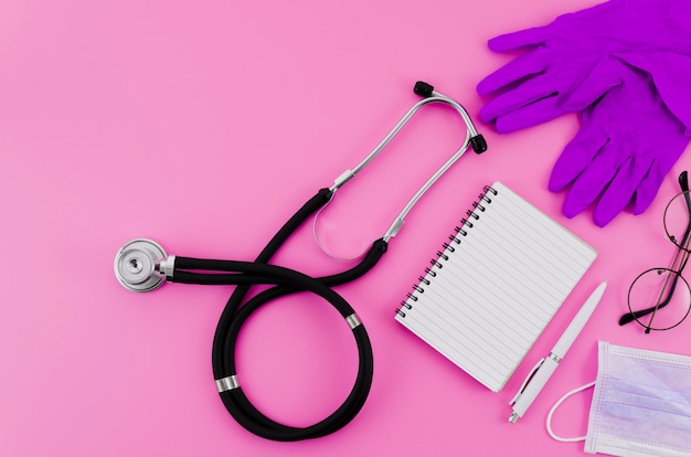 Stethoscope; surgical gloves; eyeglasses; pen; spiral notepad on pink backdrop