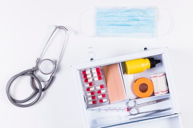 Stethoscope and surgical mask with first aid kit on white background