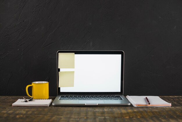 Записки на белом пустом экране ноутбука с кружкой кофе и канцелярскими принадлежностями над деревянным столом