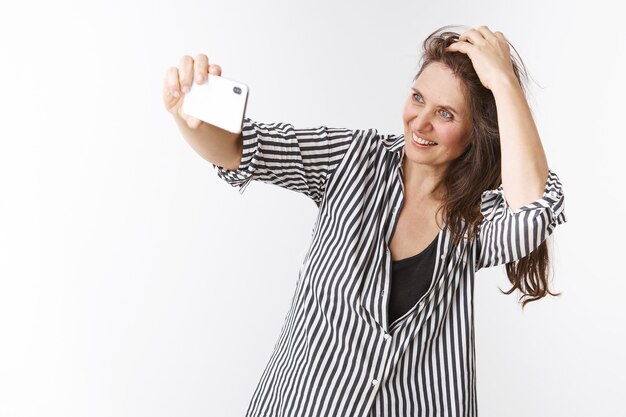 Still feeling young and pretty. Portrait of charming confident and energized middle-aged woman holding smartphone with extended hand, taking selfie checking haircut and smiling at mobile phone